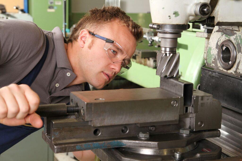 Ein Facharbeiter mit Schutzbrille überprüft die Ausrichtung eines Werkstücks an einer CNC-Fräsmaschine. Er konzentriert sich auf die präzise Positionierung des Materials während des Bearbeitungsprozesses