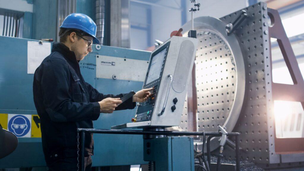 Ein Maschinenbediener mit Schutzhelm arbeitet an einer modernen CNC-Fräsmaschine in einer Industriehalle. Er steuert die Maschine über ein digitales Bedienfeld und überprüft die Einstellungen